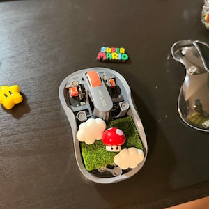 May include: A gray computer mouse with its top cover removed, revealing an internal green textured surface decorated with two white clouds and a red-and-white mushroom. The mouse features an orange scroll wheel and the text FMOUSE printed on it.