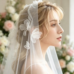 May include: A sheer white bridal veil crafted from fine tulle fabric, featuring delicate three-dimensional floral appliqués scattered across the surface. The veil is draped over the head, showcasing the translucent material and the layered petal design.