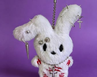 May include: A white plush rabbit keychain featuring long ears adorned with silver metal spikes, studs, and a dangling cross charm. The rabbit has black embroidered eyes and red stitched markings on its body, with a silver ball chain attached.