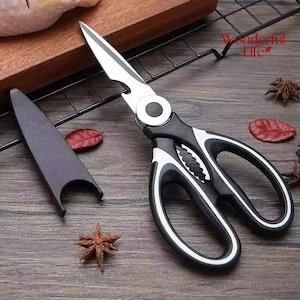May include: Multi-purpose kitchen shears with silver stainless steel blades and black ergonomic handles featuring white trim. The tool includes a built-in bottle opener and a serrated section for gripping. A matching black protective blade cover is included.