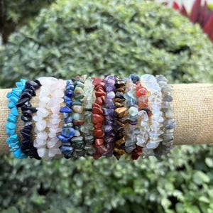 May include: A collection of eleven stretch bracelets made from various polished gemstone chips, including turquoise, lapis lazuli, rose quartz, amethyst, tiger's eye, and clear quartz, arranged in a row on a cylindrical beige fabric display stand.