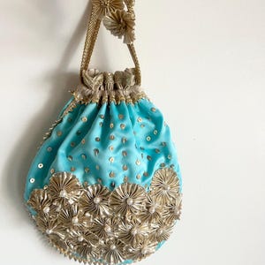Puede incluir: Un bolso tipo saco de color turquesa brillante con lentejuelas doradas esparcidas por la superficie. La parte inferior está decorada con una densa disposición de rosetas de tela dorada plisada, cada una con una pequeña perla blanca en el centro.
