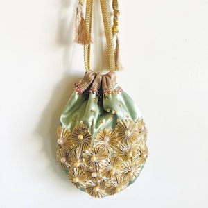 Puede incluir: Un bolso tipo saquito de satén verde salvia con cordón, decorado con adornos circulares plisados en metal dorado y pequeñas cuentas perladas. El bolso incluye asas largas de cordón trenzado dorado con borlas y lentejuelas.