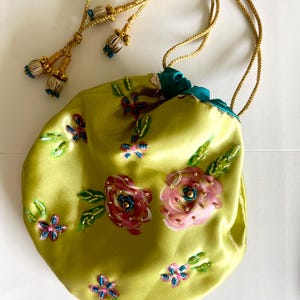 May include: A lime green satin drawstring pouch featuring hand-embroidered floral motifs in shades of pink, blue, and green. The bag includes a teal inner lining and gold-toned braided cords finished with decorative beaded tassels at the ends.
