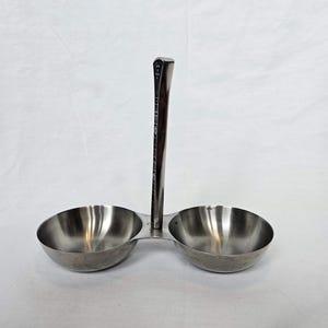May include: A stainless steel condiment server with two round bowls and a central handle. The handle is dark brown with etched markings. The bowls are shiny and appear to be empty, ready for use.