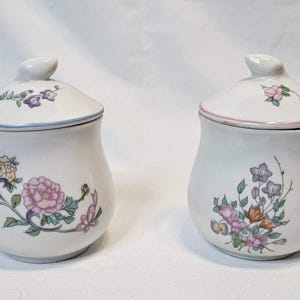 May include: Two white ceramic jars with matching lids, each featuring a unique floral pattern. The left jar displays pink, yellow, and purple flowers with green leaves, while the right jar features a cluster of purple, pink, and orange blossoms.
