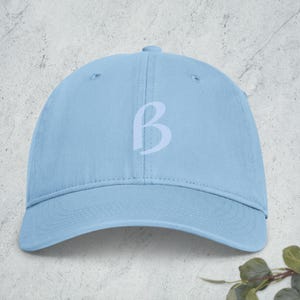 May include: A light blue baseball cap featuring a centered, embroidered uppercase letter B in a lighter shade of blue. The cap has a structured crown, a curved brim, and visible stitching details across the panels and the front bill.