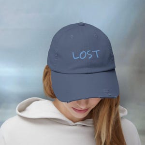 May include: A navy blue baseball cap with a distressed, worn-in finish on the brim and sides. The word LOST is embroidered in light blue lettering across the front center of the cap. The hat features a classic six-panel construction with eyelets.