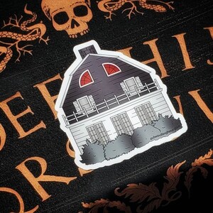 May include: A sticker featuring a black and white illustration of a house with red arched windows. The house has a dark gray roof and a white facade. The sticker is on a black surface with orange lettering and a skull.
