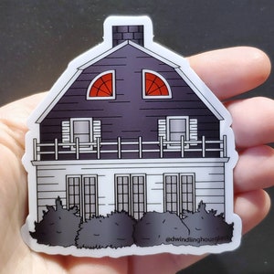 May include: A sticker featuring a two-story house with a dark purple roof and white siding. The house has two arched windows with red accents and a chimney. The bottom floor has multiple windows and a small hedge in front.