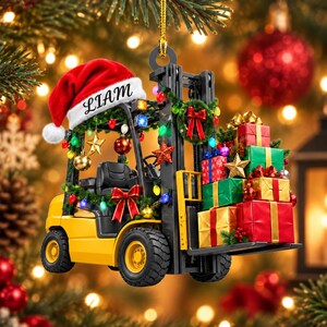 Può includere: Un addobbo natalizio a forma di carrello elevatore giallo con un cappello da Babbo Natale rosso e il nome LIAM stampato sul bordo bianco. Il carrello è decorato con ghirlande verdi, luci colorate e un fiocco rosso, trasportando pacchi regalo.