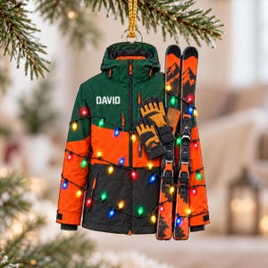 Könnte beinhalten: Ein hängender Weihnachtsbaumschmuck in Form einer Skijacke mit grünen, orangefarbenen und schwarzen Einsätzen, dem in Weiß gedruckten Namen DAVID, einem Paar orange-schwarzer Handschuhe und zwei orangefarbenen Skiern, umwickelt mit einer Lichterkette.