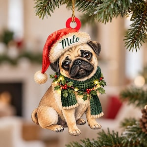 May include: A flat, hanging Christmas tree ornament featuring a tan pug dog wearing a red Santa hat with the name Milo printed on the white brim and a dark green knitted scarf adorned with holly leaves, red berries, and small glowing yellow lights.