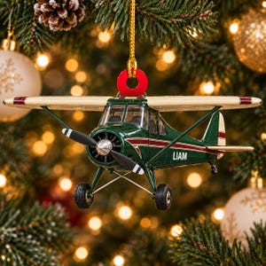 Könnte beinhalten: Ein dunkelgrünes und cremefarbenes Flugzeug-Ornament mit schwarzem Propeller und Fahrwerk. An der Seite des Rumpfes steht der Name LIAM in weißen Großbuchstaben. Eine rote kreisförmige Öse oben ist mit einer goldenen Kordel verbunden.