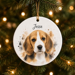 Peut inclure: Une décoration ronde en céramique blanche ornée d'une illustration style aquarelle d'un chiot beagle au pelage marron, noir et blanc. Le prénom Jesse est inscrit en lettres cursives noires au-dessus du chien. Un cordon doré est fixé.