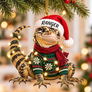 May include: A lizard ornament wearing a red and white Santa hat with the text RANGER printed on the brim, a green jumper with white snowflake patterns, and a red knitted scarf. The lizard has black and yellow scales and a long tail.