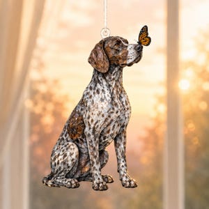 May include: A hanging ornament shaped like a German Shorthaired Pointer dog with a brown and white spotted coat. The dog is in a sitting position with a small orange and black monarch butterfly perched on its nose, suspended by a thin silver string.