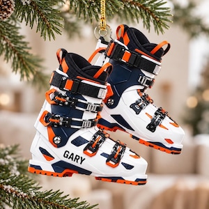 Può includere: Un paio di ornamenti a forma di scarpone da sci bianchi, blu navy e arancioni appesi a un cordino dorato. Gli scarponi presentano diverse fibbie nere e arancioni, cinturini neri e il nome GARY stampato in nero sul lato dello scafo.