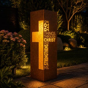 May include: A tall, rectangular, rust-colored metal garden lantern featuring a cutout cross and the text I CAN DO ALL THINGS THROUGH CHRIST WHO STRENGTHENS ME. The interior is illuminated, casting a warm glow through the cutouts in the dark.