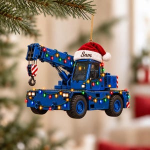 May include: A blue mobile crane Christmas tree ornament decorated with a string of multicolored LED lights. The crane features a red Santa hat on its cab with the name Sam embroidered in black script and a red and white striped hook.