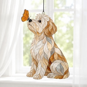 May include: A stained glass suncatcher featuring a light brown and cream-coloured dog sitting and looking up at a bright orange monarch butterfly perched on its nose. The dog is composed of individual glass segments with dark outlines.