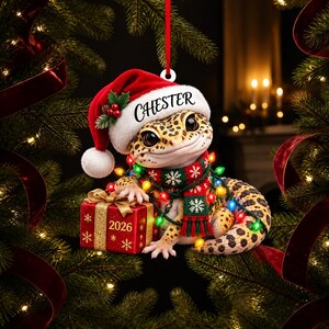 May include: A leopard gecko Christmas ornament featuring the name CHESTER on a red Santa hat, a green and red snowflake scarf, and a string of colourful lights. The gecko holds a red gift box with the year 2026 printed in gold on the front.