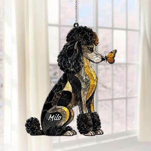Pode incluir: Um ornamento suspenso em estilo vitral em forma de cão poodle com segmentos pretos, cinzentos e dourados. O cão tem uma borboleta monarca pousada no nariz e o nome Milo impresso em branco na lateral, suspenso por uma corrente metálica.