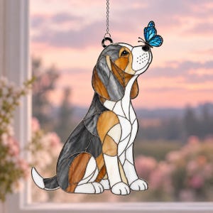 Peut inclure: Un attrape-soleil en vitrail représentant un chien beagle avec des segments noirs, marron et blancs. Le chien est assis et regarde un papillon bleu vif posé sur son museau. L'objet est suspendu par une chaîne et un anneau en métal.