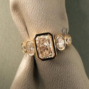 Könnte beinhalten: Ein goldener Ring mit einem großen, rechteckigen, hellfarbigen Edelstein mit schwarzem Rand, flankiert von kleineren, runden Edelsteinen. Der Ring wird vor einem neutralfarbenen Stoff präsentiert.