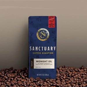 May include: A navy blue coffee bag with gold lettering and a gold emblem. The bag reads "SANCTUARY COFFEE ROASTING" and "MIDNIGHT OIL". The bag is surrounded by roasted coffee beans. The bag contains 12 oz (340 g) of coffee.