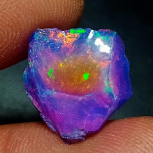 May include: A raw, unpolished opal gemstone featuring a vibrant play of color with deep purple and blue base tones, accented by bright flashes of orange, yellow, and neon green throughout its irregular, multifaceted surface structure.