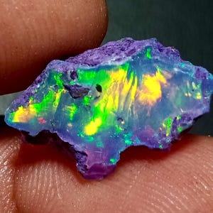 Op de afbeelding: Een ruwe, ongepolijste opaal edelsteen met een doorschijnende blauwe basis en levendige flitsen van neongroen en heldergeel kleurenspel. De steen heeft een onregelmatige, grillige vorm met een ruwe, getextureerde paarse rand.
