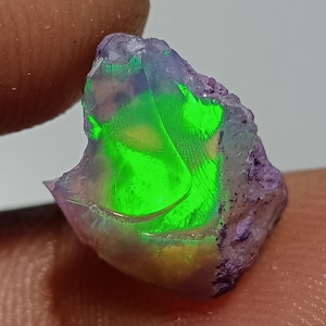 Op de afbeelding: Een ruwe, ongeslepen opaal edelsteen met een doorschijnende, onregelmatige vorm en een levendig, gloeiend neon-groen kleurenspel in het midden, gecontrasteerd door ondoorzichtige, getextureerde paarse en lavendelkleurige minerale randen.