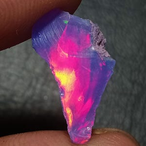 Op de afbeelding: Een ruw, doorschijnend opaal edelsteenfragment met een levendig kleurenspel met intense flitsen van neonroze, felgeel en elektrisch blauw tegen een halfdoorzichtige, lichtviolette basis met een ruwe, ongepolijste randtextuur.