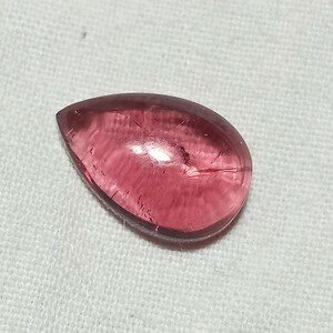 May include: A pear-shaped, translucent gemstone with a deep pinkish-red hue. The stone features a smooth, polished surface with visible internal inclusions and natural striations that catch the light, creating a subtle glow across its faceted body.
