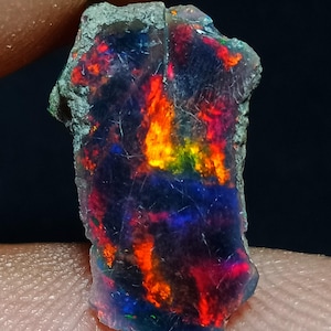 May include: A raw, uncut black opal gemstone featuring a dark, translucent base with vibrant flashes of red, orange, and green play-of-colour. The stone has an irregular, jagged shape with a rough, unpolished texture along the outer edges.