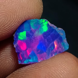 Op de afbeelding: Een ruwe, ongepolijste opaal edelsteen met een levendig kleurenspel met intense flitsen van neonblauw, elektrisch groen en helder magenta. De steen heeft een onregelmatige, grillige vorm met een doorschijnende, veelzijdige oppervlaktestructuur.