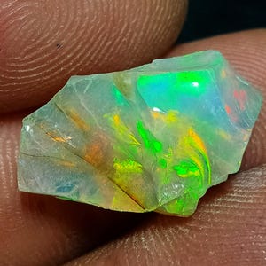 May include: A raw, translucent opal gemstone featuring a play-of-color with vibrant flashes of neon green, bright orange, and yellow against a pale, milky white base. The stone has an irregular, angular shape with visible natural surface fractures.
