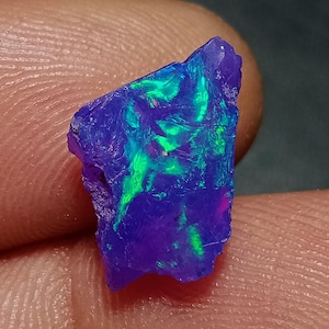 May include: A raw, irregularly shaped opal gemstone featuring a deep purple base color with vibrant, iridescent flashes of bright neon green and subtle hints of pink throughout its textured, crystalline surface, showcasing natural geological patterns.
