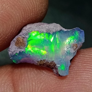 Op de afbeelding: Een ruwe, ongepolijste opaal edelsteen met een doorschijnende, melkblauwe basis met levendige, iriserende flitsen van neongroen en geel licht. De steen heeft een onregelmatige, grillige vorm met vlekken van ruw, bruingrijs gesteente.