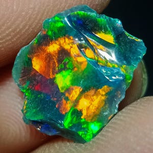 Puede incluir: Una piedra preciosa de ópalo en bruto y sin tallar con un color base azul profundo translúcido y destellos multicolores vibrantes de verde neón, naranja brillante y amarillo en toda su superficie irregular y dentada.