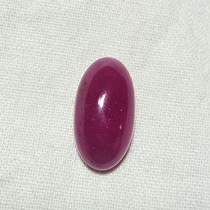 May include: An oval-shaped, deep magenta-colored gemstone with a smooth, polished cabochon surface. The stone features a glossy finish that reflects light, highlighting its solid, opaque, and uniform dark pinkish-red color throughout the piece.