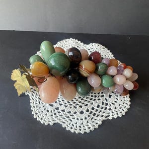 May include: A decorative cluster of polished stone grapes in various shades of green, pink, brown, and translucent white. The grapes are arranged in a bunch with a metallic gold-colored leaf attached to the stem on the left side of the object.