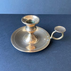 May include: A brass candlestick holder with a circular base and a small handle. The candle holder has a cup-shaped top to hold a candle. The metal has a warm, golden hue.