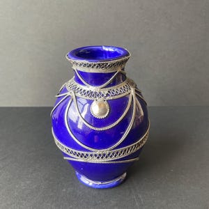 Può includere: Un vaso blu cobalto con dettagli in filigrana d'argento. Il vaso ha un corpo arrotondato e un collo stretto. La filigrana d'argento è disposta in motivi decorativi attorno al vaso, con un piccolo disco d'argento al centro.