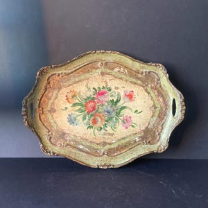 Könnte beinhalten: Ein ovales Serviertablett mit einem abgenutzten hellgrünen und goldenen Finish. Die Mitte zeigt einen handgemalten Blumenstrauß mit roten, rosa, orangefarbenen und blauen Blüten auf cremefarbenem Grund. Das Tablett hat zwei integrierte Griffe.