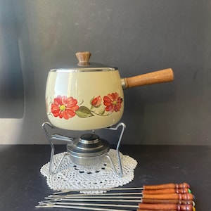 Puede incluir: Una olla para fondue de color crema con un patrón floral de flores rojas y hojas verdes, que cuenta con un mango y un pomo de tapa de madera. Se asienta sobre un soporte de metal con quemador. Se incluyen seis tenedores de fondue con mangos de madera.