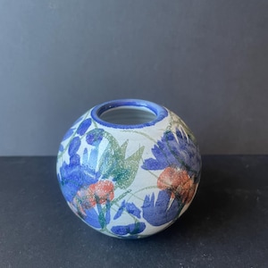 Può includere: Un vaso in ceramica sferico con base bianca caratterizzato da un motivo floreale dipinto a mano nei toni del blu, verde e arancione. Il vaso presenta un'apertura circolare sulla parte superiore con un bordo blu e un corpo liscio e arrotondato.