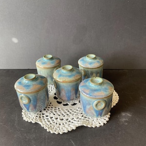 Op de afbeelding: Een set van vier kleine keramische potten met deksel, voorzien van een reactief glazuur in blauwe, beige en bruine tinten. Elke cilindrische pot heeft een bijpassend deksel met een kleine knop en een cirkelvormig decoratief accent op de zijkant.