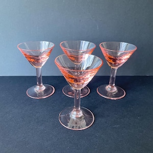 Puede incluir: Cuatro copas de cóctel rosas con un cuenco transparente y un borde y un tallo rosas. Las copas están dispuestas sobre una superficie oscura. Las copas tienen una forma clásica de copa de Martini.